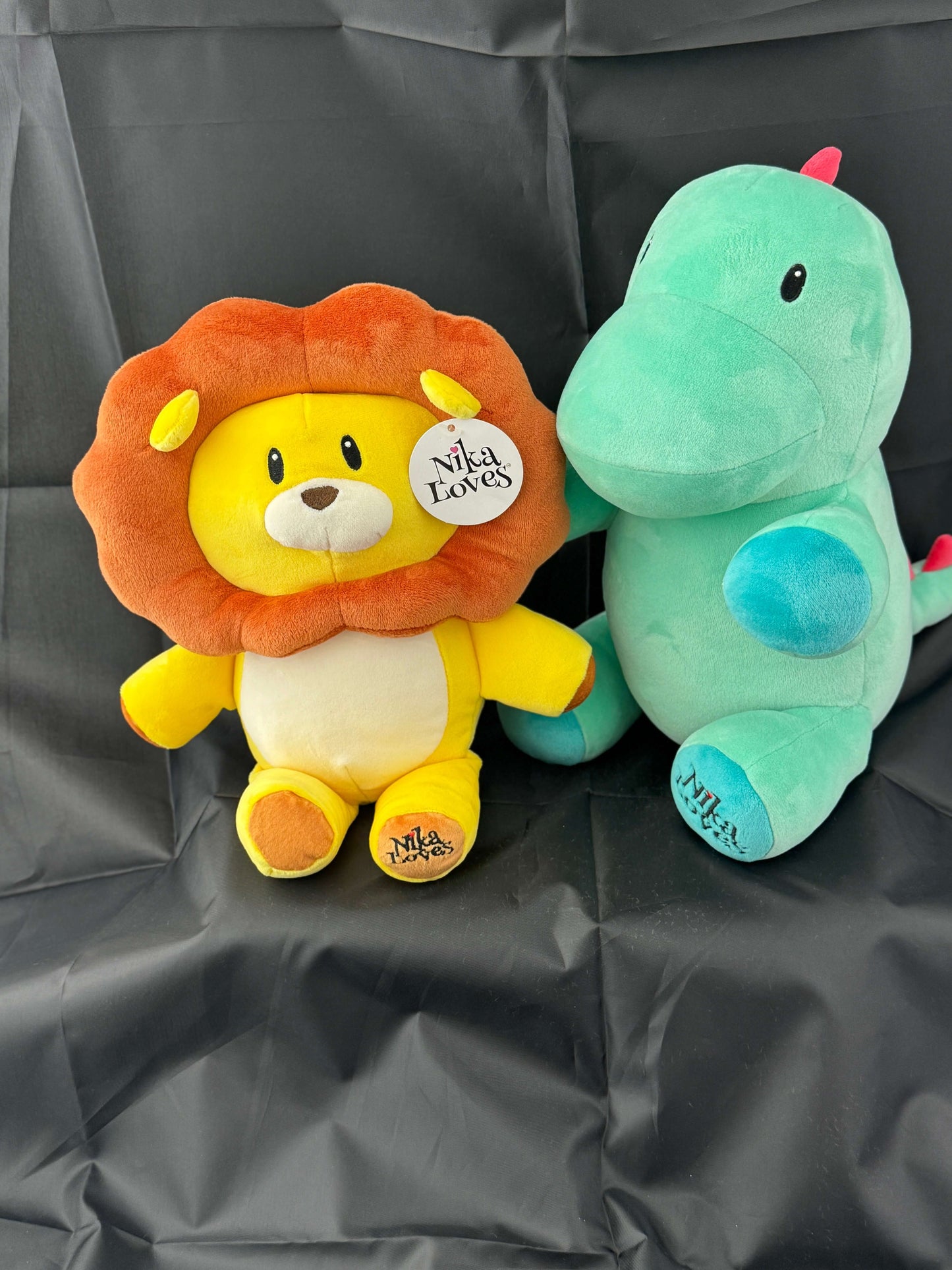 Two plush toys, a lion and a green dinosaur, with "Nila Loves" tags, sitting on a black fabric backdrop.