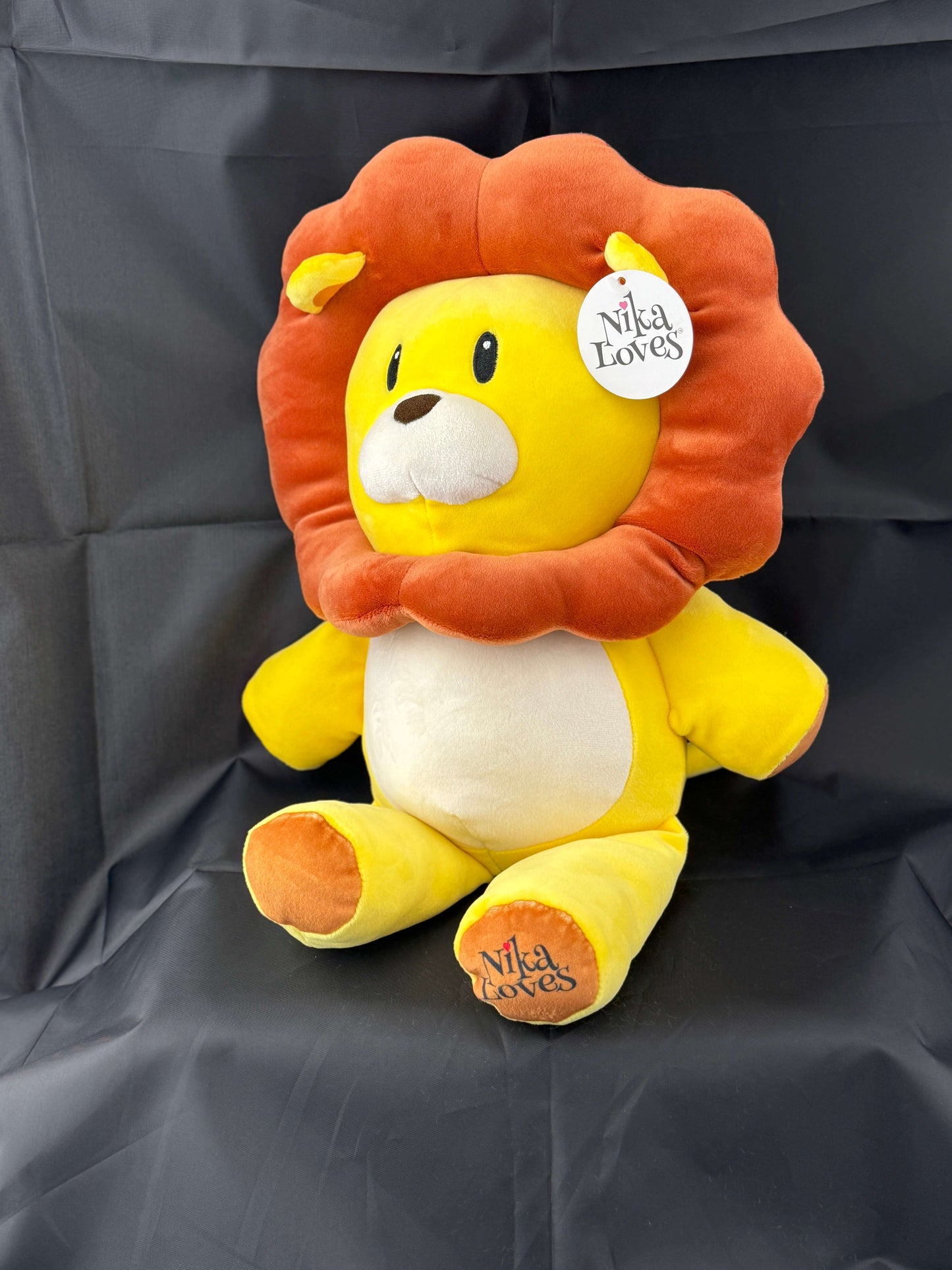 Plush lion toy with orange mane and "Nila Loves" tags sitting on a black surface.