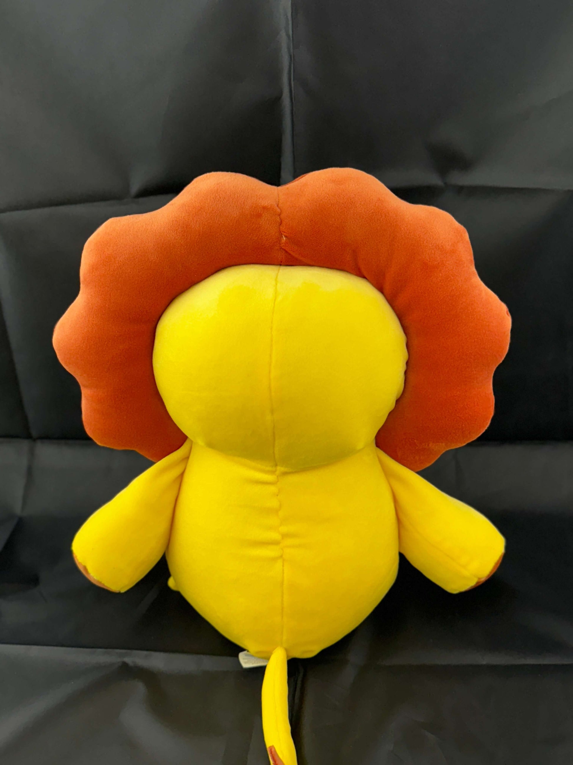 Plush stuffed lion toy with a yellow body and orange mane, sitting on a black background.