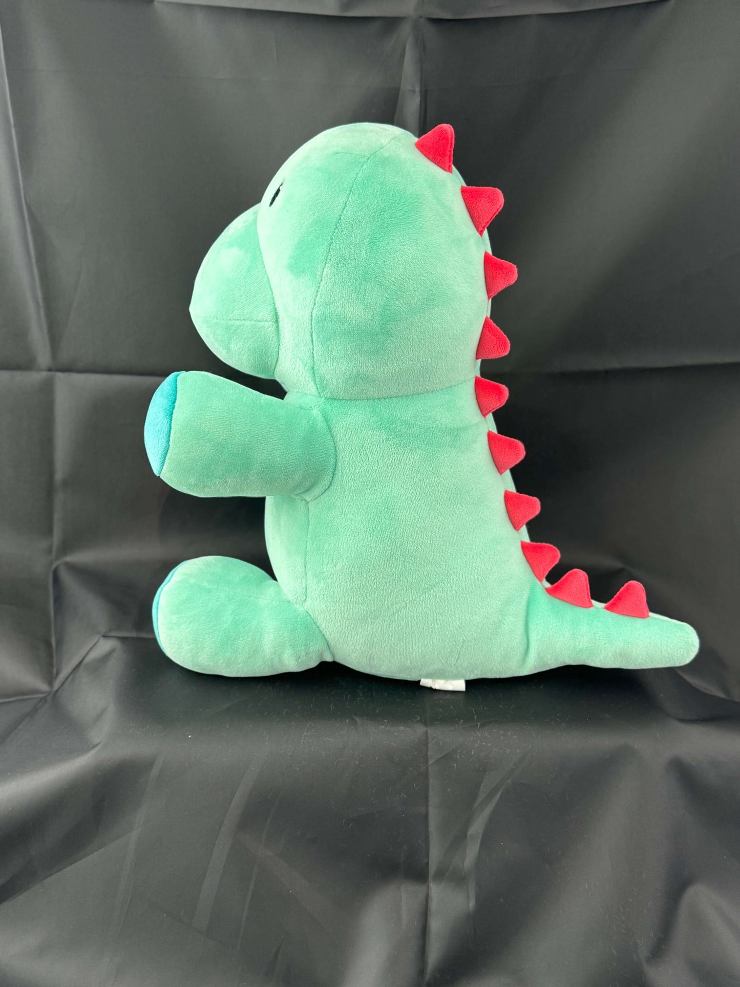 Green Genosaurus plush dinosaur toy with red spikes and a blue belly, sitting on a black background.